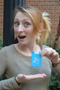 This image has a smiling woman with strawberrt blond hair and freckles showing off a Carolina Blue 2-step credit card pocket that you can attach to your phone.